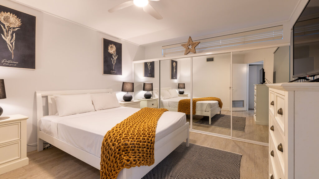 A cozy modern bedroom with a white bed frame, white bedding, and a chunky mustard knit throw, flanked by matching nightstands and lamps, with mirrored closet doors, botanical artwork above the bed, and light wood flooring with a striped rug.
